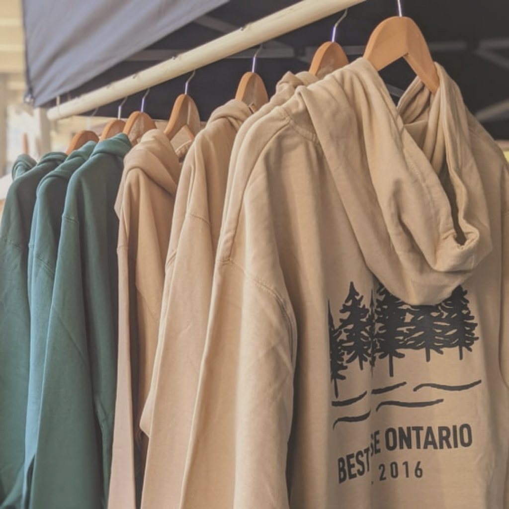 Beige and forest green hoodies hanging on a rack with a black logo across the shoulders. Logo has six tree and five waves and says "Best Case Ontario est 2016" 